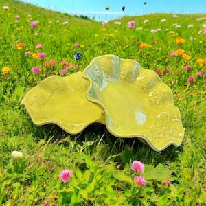 May include: Two yellow ceramic bowls with a wavy edge and floral design, sitting on a bed of green grass with pink and yellow flowers.