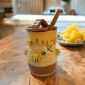 May include: A ceramic honey pot with a lid and a honey dipper. The pot is yellow with a honeycomb pattern and bee accents. The lid and dipper are brown. A plate of honeycomb is in the background.