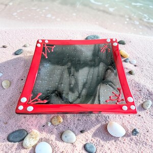 May include: A square, red-framed glass dish with a black and gray interior. Coral-like designs in red adorn the corners. White dots are placed on the corners. The dish is resting on pink sand with small rocks and seashells.