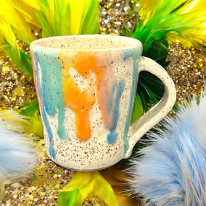 May include: A white ceramic mug with a speckled glaze and a rainbow drip design. The mug has a rounded handle.