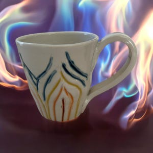 May include: A handmade ceramic mug with a white base and a curved handle. The mug features a design of vertical lines in blue, yellow, and orange. The mug is a unique piece of pottery.