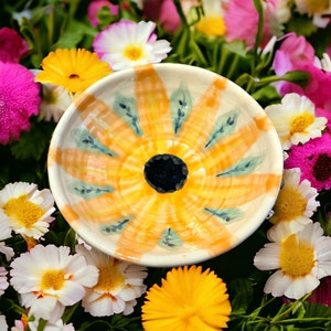 May include: A small, white ceramic bowl with a yellow and orange floral design. The bowl has a raised rim and a smooth, glazed finish.