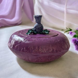 May include: A purple ceramic trinket box with a lid, featuring embossed thistle designs. The lid is topped with a dark blue thistle sculpture and green leaves. The box is a vibrant shade of purple.