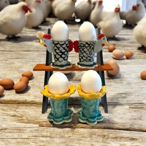 May include: Two sets of ceramic egg cups, one set with a chicken design and the other with a floral design. The chicken egg cups are gray with black accents and red combs. The floral egg cups are turquoise with yellow accents. Each set has two egg cups.
