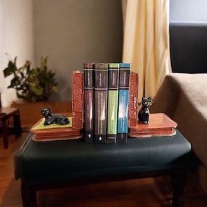 May include: A set of brown bookends with black cat figurines on top. The bookends are holding up a row of five books with green, blue, and black spines. The text on the spines reads "The Collected Writings of Franz Liszt" and "The Collected Writings of Franz Liszt, Volume 2".