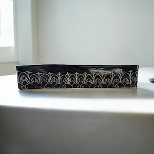 May include: A rectangular black ceramic dish with a white floral pattern along the top edge. The dish is sitting on a white surface, and the design is symmetrical.