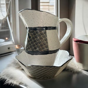 May include: A ceramic pitcher and matching bowl set. The pitcher has a large handle and a speckled white finish with a black band and fan-shaped pattern. The bowl has a scalloped edge and a similar fan pattern.
