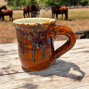 May include: A brown ceramic mug with a white rim and a unique handle. The mug is sitting on a wooden table with a blurred background of horses in a field.