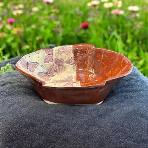 May include: A small, handmade ceramic dish with a brown glaze. One side of the dish is decorated with a white background and a pattern of pink butterflies. The other side of the dish is a solid brown color.