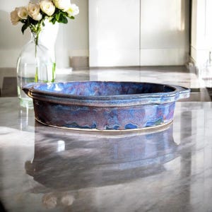 May include: A handmade, oval-shaped ceramic baking dish with a blue and purple glaze. The dish has a slightly raised rim and two small handles. The background includes a vase of white roses and a marble countertop.