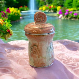 May include: A pink and white ceramic jar with a lid. The lid has a swirl design on top. The jar is sitting on a pink tablecloth.