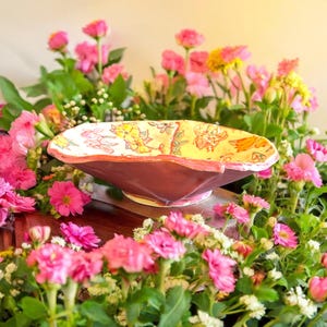 May include: A hand-painted ceramic bowl with a pink and yellow floral design. The bowl is sitting on a wooden surface surrounded by pink flowers.