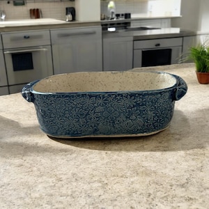 May include: A blue ceramic baking dish with a floral pattern. The rectangular dish has rounded corners and handles on each end. The interior is speckled with a light beige color.