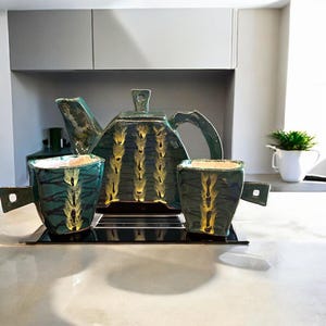 May include: A ceramic tea set in shades of teal and gold. The set includes a geometric teapot and two square-shaped cups with handles. The teapot and cups have a textured design with vertical gold accents. The set is displayed on a black tray.