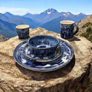 May include: A blue and white ceramic dinnerware set with a bowl, plate, and cup. The set is on a rock with a mountain range in the background.