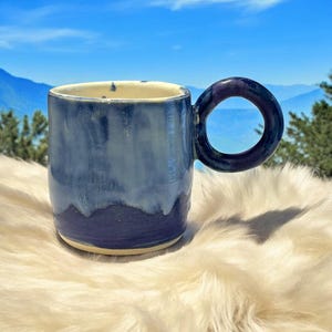 May include: A ceramic mug with a light blue and purple glaze. The mug has a unique, rounded handle.