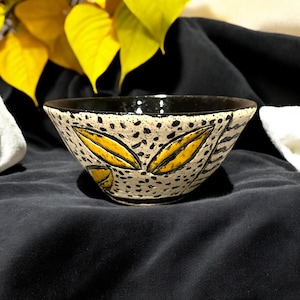 May include: A small ceramic bowl with a black rim and a white interior. The bowl is decorated with black and yellow geometric patterns.