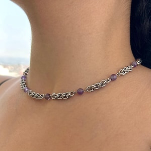 Peut inclure: Un collier en chaîne argenté orné de petites perles rondes violettes. Le collier est porté autour du cou et la chaîne présente un motif tissé. Le collier est en métal.