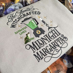 May include: Embroidered beige fabric with the text "The Witchcrafted Midnight Margaritas." The design includes a blender with green liquid, a frog, and a candle. The words "Barbados Limes" are also present.