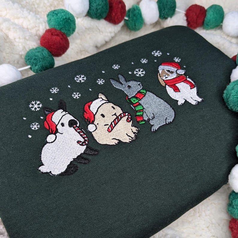 May include: Dark green sweatshirt featuring an embroidered design of four rabbits in festive attire. Each rabbit is wearing a Santa hat or scarf, with snowflakes scattered around them. A candy cane is held by one rabbit. A Christmas-themed design.