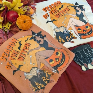 Könnte beinhalten: Zwei T-Shirts mit Halloween-Motiv. Jedes Shirt zeigt eine Hexen-Grafik, die auf einem geschnitzten Kürbis sitzt, mit dem Text "Season of the Witch". Die Shirts sind in Orange- und Cremetönen gehalten, mit schwarzen und orangen Grafiken. Ein dekoratives schwarz-weißes Objekt ist ebenfalls sichtbar.