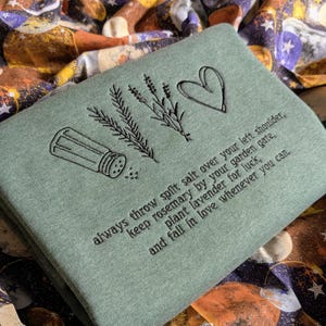 May include: Sage green sweatshirt with embroidered black line art of a salt shaker, sprigs of rosemary and lavender, and a heart. Text reads: "always throw spilt salt over your left shoulder...and fall in love whenever you can."