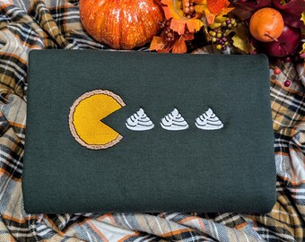 Può includere: Felpa verde scuro con un design ricamato raffigurante una fetta di torta gialla e tre ciuffi di panna montata bianca. La felpa è esposta su una coperta a quadri con una zucca e foglie autunnali sullo sfondo.