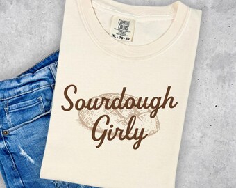 May include: A beige t-shirt with the text "Sourdough Girly" in brown lettering. A brown illustration of a loaf of sourdough bread is above the text.