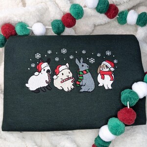 May include: Dark green sweatshirt featuring an embroidered design of four rabbits wearing Santa hats and scarves, with snowflakes. Two rabbits hold candy canes. A festive pom-pom garland in red, green, and white decorates the background.