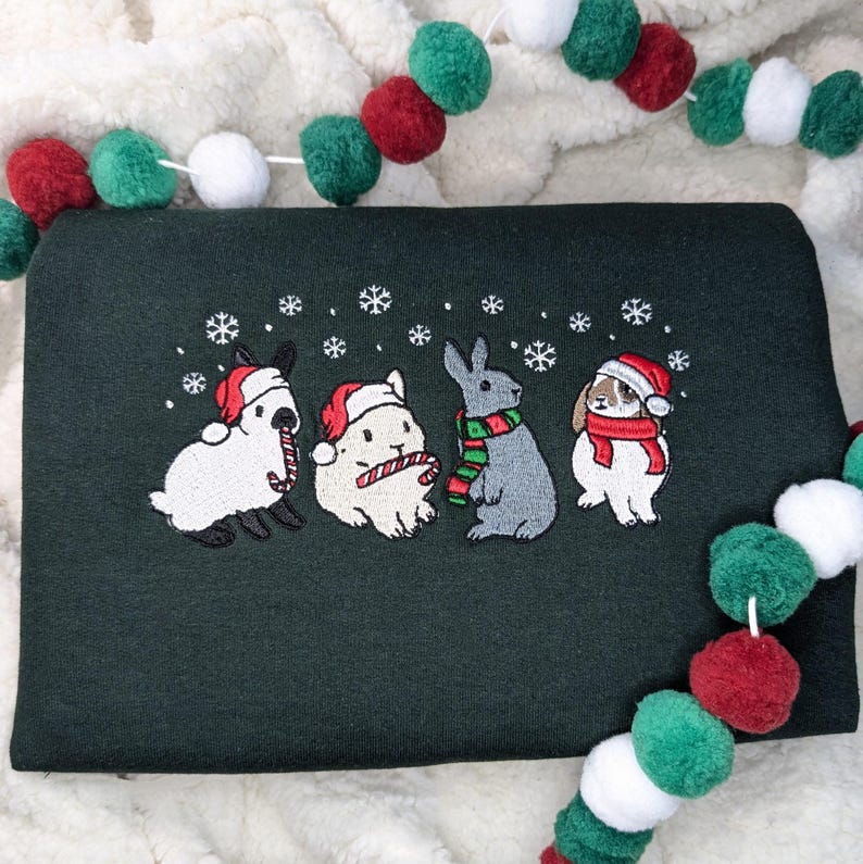 May include: Dark green sweatshirt featuring an embroidered design of four rabbits wearing Santa hats and scarves, with snowflakes. Two rabbits hold candy canes. A festive pom-pom garland in red, green, and white decorates the background.