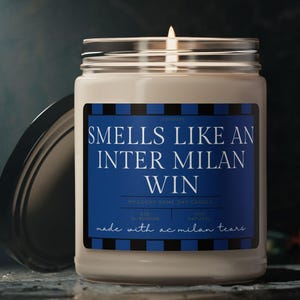 May include: A lit scented candle in a glass jar with a silver lid. The label reads "Smells Like An Inter Milan Win" in white text on a blue background with black and blue stripes. The text "made with ac milan tears" is also on the label.