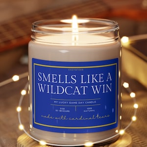 May include: A brown candle with a blue label that reads "Smells Like A Wildcat Win" and "My Lucky Game Day Candle". The candle is on a wooden coaster and surrounded by white fairy lights.
