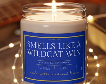 May include: A brown candle with a blue label that reads "Smells Like A Wildcat Win" and "My Lucky Game Day Candle". The candle is on a wooden coaster and surrounded by white string lights.