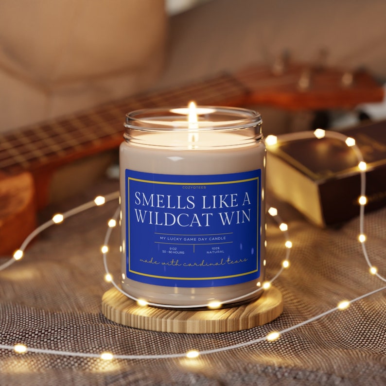 May include: A brown candle with a blue label that reads "Smells Like A Wildcat Win" and "My Lucky Game Day Candle". The candle is on a wooden coaster and surrounded by white fairy lights.