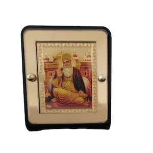 May include: A gold-coloured framed image depicting a religious figure with a white beard and yellow turban, seated in meditation. The background shows a golden temple. The frame has a black border and silver studs.