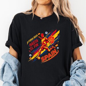 May include: A black t-shirt featuring a graphic of a soccer player in red and yellow, with the text "FURIA ROJA", "HASTA EL FIN", and "SPAIN". The design includes diagonal red and yellow brush strokes, soccer balls, and the Spanish national crest.