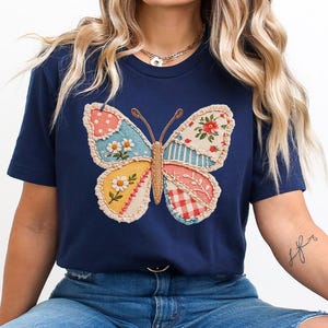 May include: Navy blue t-shirt featuring a colorful butterfly design. The butterfly has patchwork wings with floral, polka dot, and striped patterns. The shirt is a casual, everyday wear item.