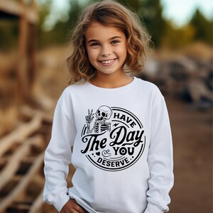 May include: White long-sleeve sweatshirt featuring a black graphic of a skeleton giving a peace sign. The text "Have The Day You Deserve" is printed in a circular design. The sweatshirt is designed for children.