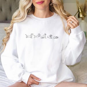 Peut inclure: Sweat-shirt blanc avec une ligne de cinq teckels en contour noir, tous face &agrave; droite de l'image.