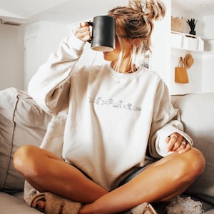 Peut inclure: Une femme portant un sweat-shirt blanc avec une ligne de silhouettes de teckels noires. Le sweat-shirt a un col rond et des manches longues. La femme est assise sur un canap&eacute; et tient une tasse noire.