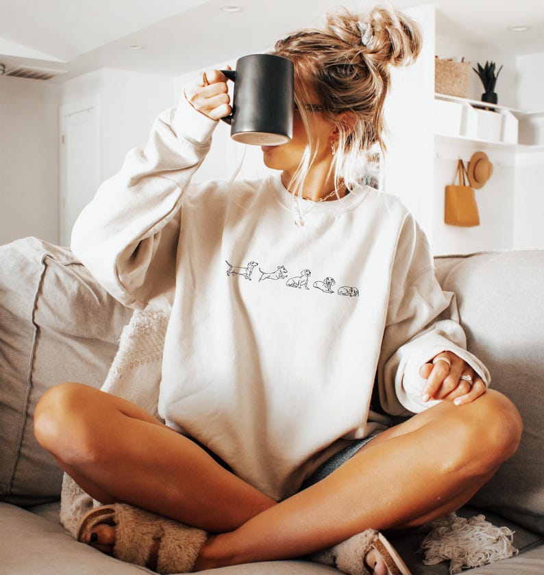 Peut inclure: Une femme portant un sweat-shirt blanc avec une ligne de silhouettes de teckels noires. Le sweat-shirt a un col rond et des manches longues. La femme est assise sur un canap&eacute; et tient une tasse noire.