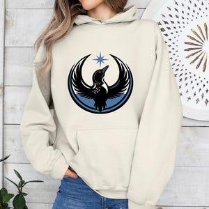 May include: Cream-colored hoodie featuring a graphic of a black bird with outstretched wings, a red eye, and a blue star. The bird is encircled by a blue and black ring. The hoodie has a front pocket and a drawstring hood.