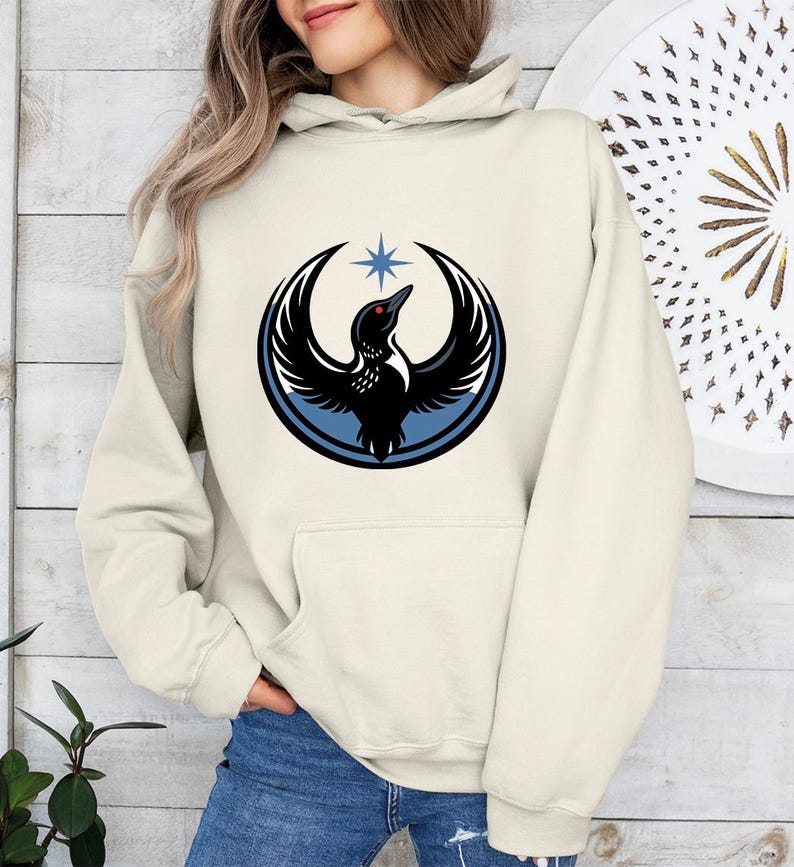 May include: Cream-colored hoodie featuring a graphic of a black bird with outstretched wings, a red eye, and a blue star. The bird is encircled by a blue and black ring. The hoodie has a front pocket and a drawstring hood.