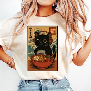 May include: Cream-colored t-shirt featuring a graphic of a black cat wearing headphones and holding a game controller. The cat is in front of a bowl of ramen with Japanese text and a wave design in the background. The design is in a rectangular frame.