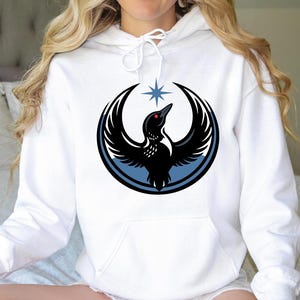 May include: White hoodie with a large graphic of a black bird with outstretched wings, a red eye, and a star above it. The bird is inside a black circle with a blue border. The hoodie has a front pocket and a drawstring hood.