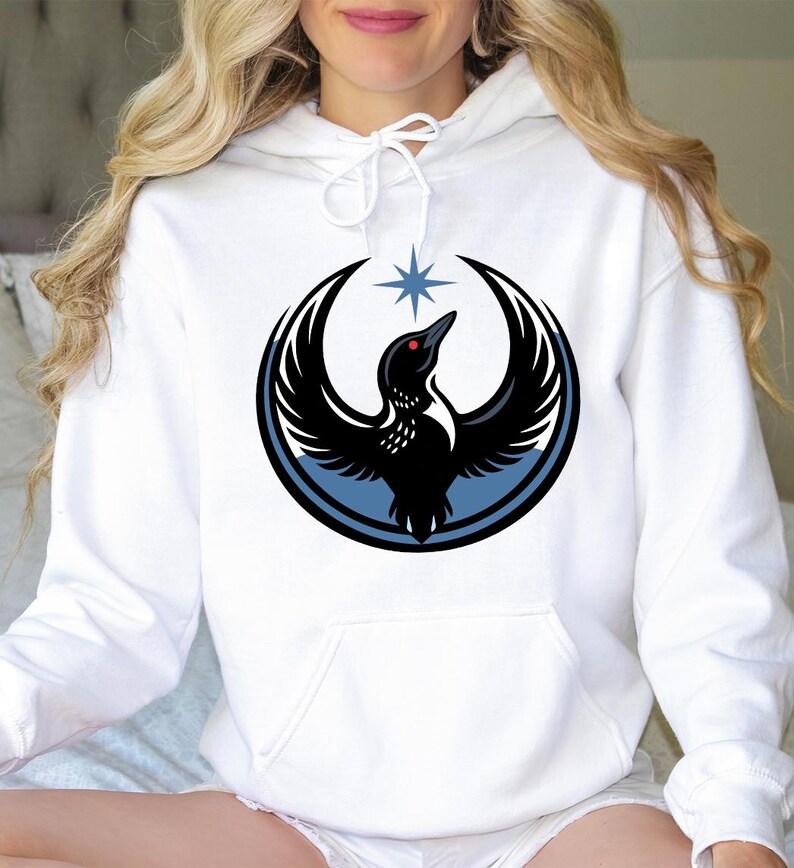 May include: White hoodie with a large graphic of a black bird with outstretched wings, a red eye, and a star above it. The bird is inside a black circle with a blue border. The hoodie has a front pocket and a drawstring hood.