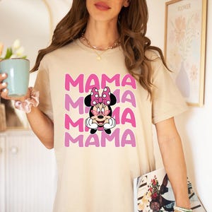 May include: A beige t-shirt with a pink and white graphic of Mickey Mouse's girlfriend Minnie Mouse. The graphic says "MAMA" four times in a stacked design.