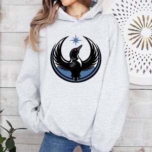 May include: A light gray hoodie featuring a black bird emblem with outstretched wings, a red eye, and a blue star above. The bird is encircled by a blue and black design. The hoodie has a drawstring hood and a front pocket.