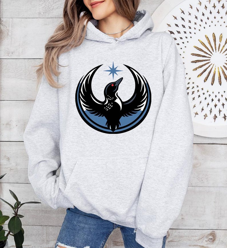 May include: A light gray hoodie featuring a black bird emblem with outstretched wings, a red eye, and a blue star above. The bird is encircled by a blue and black design. The hoodie has a drawstring hood and a front pocket.