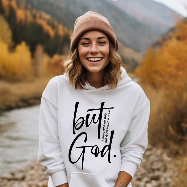 May include: White hoodie with the text "but God" in black script. The hoodie has a front pocket and a drawstring hood. The person in the image is wearing a brown beanie. The background is a blurred outdoor scene with trees.